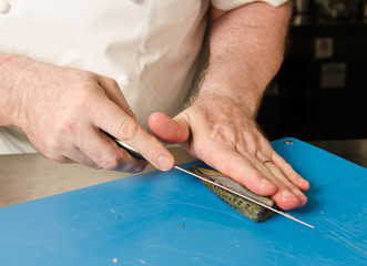 A knife, being held by a male chefs hands, slicing through fresh mackerel fish fillet, on a blue chopping board. mackerel is high in omega fish oils.