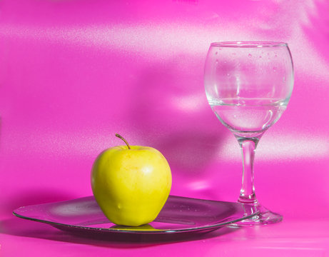 on a pink background on the plate yellow Apple with a glass of water.