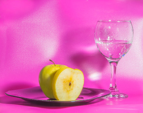 on the plate is yellow Apple with a glass of water on a pink background.