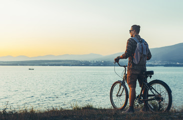 Fototapeta na wymiar Young Man Cyclist Standing With Bike On Coast And Enjoying View Of Sunset Vacation Traveling Destination Resting Concept