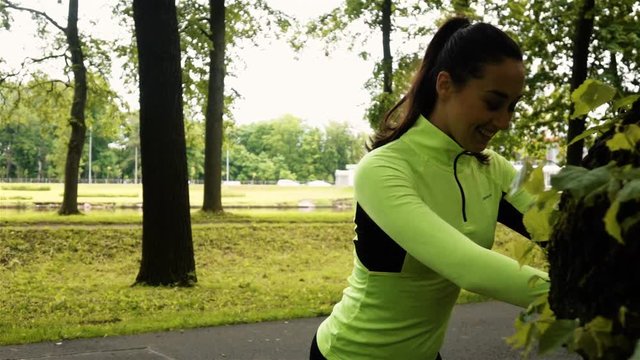 Young sexy woman exercising in park near tree with asphalt road. She smiling and waving leg. Super slow motion.