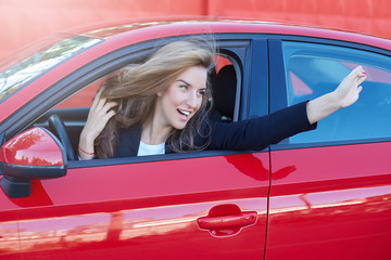 Portrait of young and beautiful Caucasian business lady with long hair wearing formal suit on the wheel of red modern car hanging and flirting with her college while parking at the office.