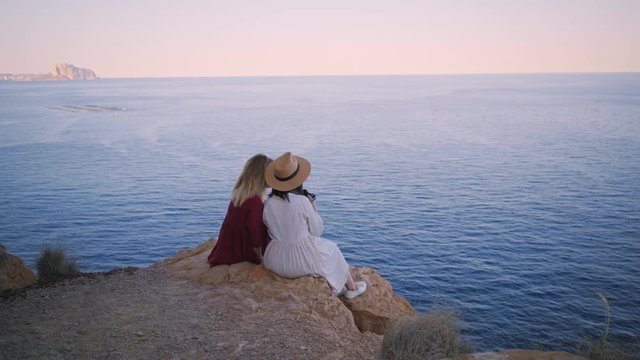 Static shot of two girls watching beautiful landscape with calm sea and warm purple sunset, they sit on rock next to each other, make photos and laugh, concept summer, friendship, youth and adventure