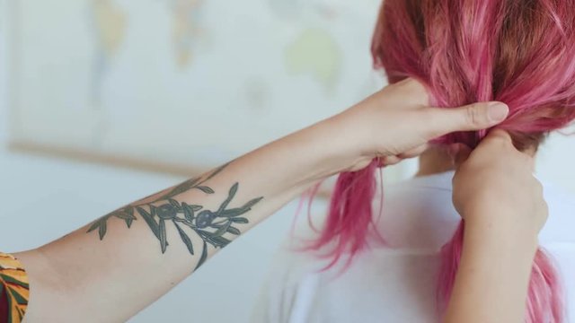 Hand held shot of young authentic hipster millennial girl with tattoos doing a braid to her friend a young woman with rose pink unconventional hair style, concept friendship, special and original