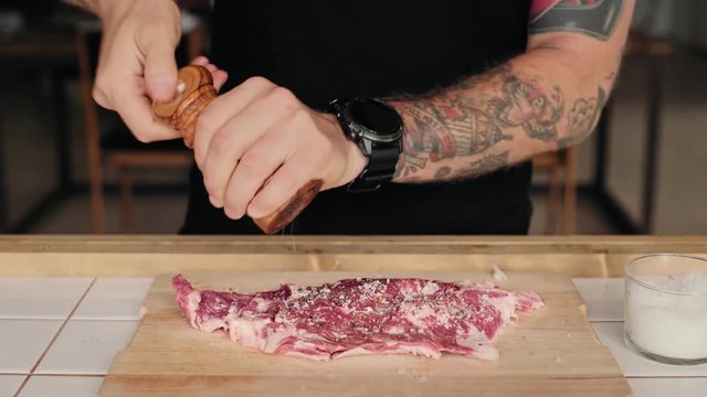 Confident independent casual hipster man with arm tattoos prepares big chunk of fresh organic farm grown meat to oven or grill, seasons and marinates it in sea salt and grounded pepper