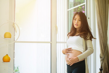 Beautiful pregnant woman is touching her belly and smiling while standing near the window at home