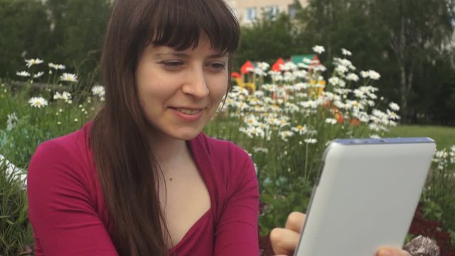Portrait of young attractive brunette woman with long hair using a white tablet computer sitting next to daisies in the city park. The girl smiles reading messages on the touch screen on a summer day.