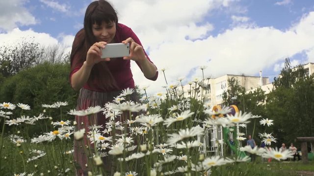 Young happy brunette woman in pink blouse and colorful skirt photographed with white phone blooming daisies in city park on a summer day.