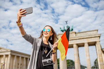 Fototapeta premium Młoda kobieta turysta robi selfie zdjęcie z niemiecką flagą stojącą przed słynną bramą Brandenburgii w Berlinie