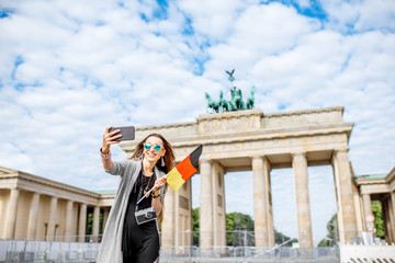 Fototapeta premium Young woman tourist making selfie photo with german flag standing in front of the famous Brandenburg gates in Berlin