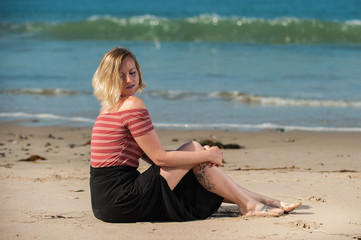 Fototapeta na wymiar Beautiful short hair woman seated on beach sand peeking over shoulder and caressing knee. 