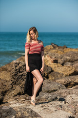 Lovely blond in long black skirt standing at rock jetty at California beach.