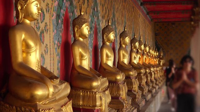 Worshipers walk past row of golden buddha statues at Wat Arun temple in Bangkok, Thailand (faces blurred for commercial use)