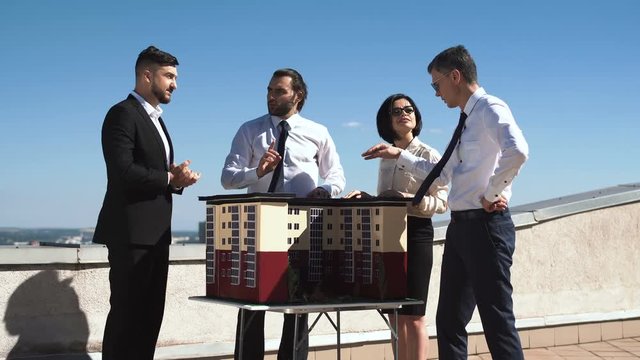 Group of engineers and architects in a meeting standing outdoors on a pation discussing a model of office or apartment blocks in a low angle view against blue sky.