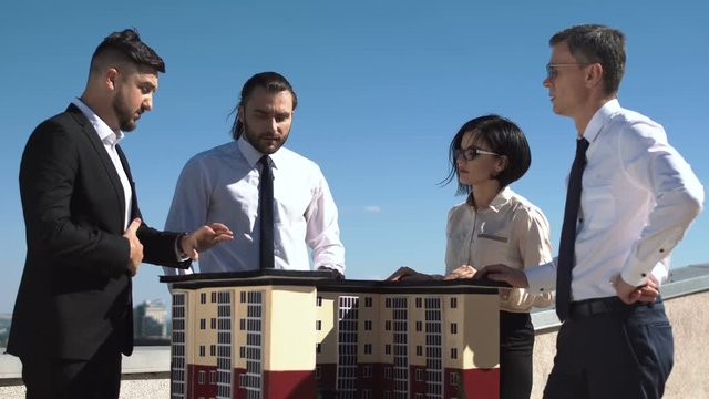 Group of engineers and architects in a meeting standing outdoors on a pation discussing a model of office or apartment blocks in a low angle view against blue sky.