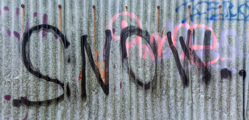 Detail of dirty corrugated iron with sprayed snow lettering. Wavy texture of urban fence from metal sheet with black, pink and blue writing. Idea of graffiti, youth culture or vandalism.