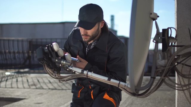 Engineer or technician checking or installing a parabolic dish television antennae and booster on the roof of an urban high-rise using digital tablet