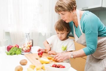 Happy grandmother and her little grandson making smoothie with fresh fruit and berries in the blender in kitchen. Little  boy preparing fruit cocktail or smoothie for breakfast. Healthy eating.