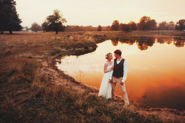 The bride and groom posing against the background of the lake in the red sunset. Overall plan
