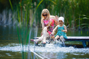 Mom and daughter are sitting on the bridge near the lake and splashing their feet
