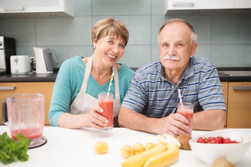 Happy seniors holding glasses with healthy homemade fruit drink fresh smoothie in kitchen
