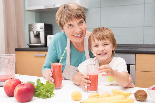 Happy grandmother and her little grandson drink smoothie with fresh fruit and berries preparing in kitchen. Little  boy preparing fruit cocktail or smoothie for breakfast. Healthy eating