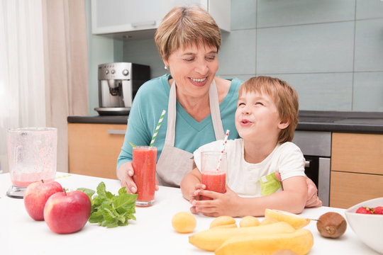 Happy grandmother and her little grandson drink smoothie with fresh fruit and berries preparing in kitchen. Little  boy preparing fruit cocktail or smoothie for breakfast. Healthy eating