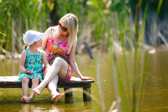 Mother and daughter sits on a wooden bridge with feet in the water