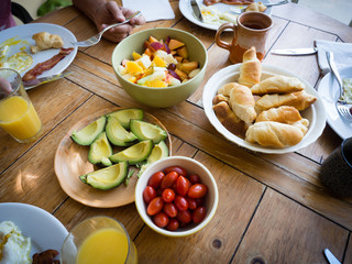 Healthy American breakfast with fruit salad, tomatoes, avocado, eggs, bacon, bread, orange juice on farm style wood table
