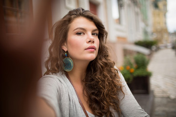 Travelgirl with long curly hair doing selfie in pedestrian street. Pretty woman looking at camera and taking selfie shot.