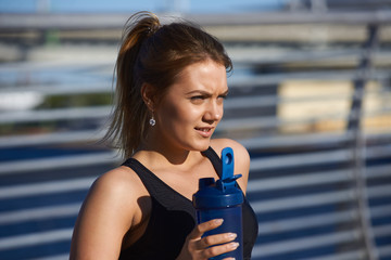 Tired thirsty woman runner with ponytail drinking water after running workout, catching her breath and relaxing. Pretty girl having break during cardio training outdoors, enjoying fresh drink