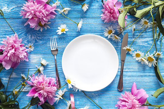 Festive table setting with cutlery, peonies and daisies on blue wooden background. Top view.