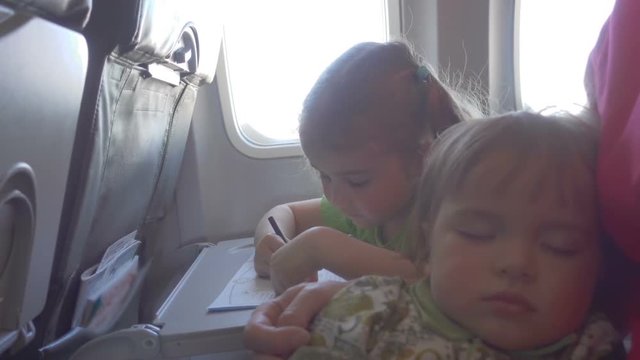 Little girl child draws,  sitting on the board of plane. Baby boy is sleeping with mom in her arms.