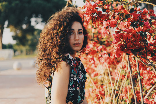 Beautiful caucasian female wearing colorful summer dress is looking at the camera while standing beside the flowers on a summer day. Brunette hipster girl with long curly hair is having rest outdoors.
