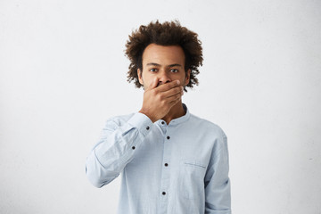 Dark-skinned mixed raced male in formal wear covering his mouth trying to keep silence and to tell secret. Tired student male yelling while wanting to sleep after sleepless night keeping hand on mouth