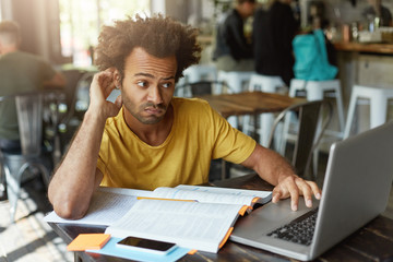 Stylish student with African hairstyle wearing casual clothes having doubtful look while looking at...