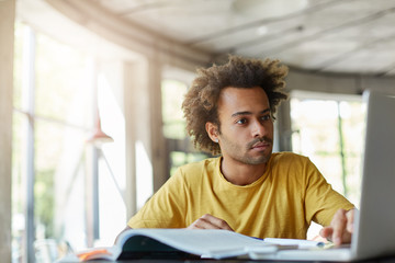 Stylish Afro American hipster male with bushy hairstyle wearing casual T-shirt being focused into screen of laptop sitting in spacious light room with big windows working with literature and internet