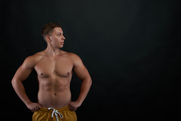 Fototapeta na wymiar Front view of young man look to away to the copy space to advertising sport product. Fit male model standing with his hands on hips against black background. Muscular and athletic guy body