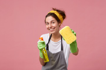 Smiling housewoman in apron and rubber gloves showing tidy sponge and detergent isolated over pink background with copy space. Beautiful joyful maid going to tidy up her room with washing spray