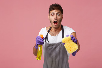 Untidy janitor wearing apron and rubber gloves looking with bugged eyes and jaw dropped while doing house cleaning with sponge and detergent having surprised expression to see much dust and dirt