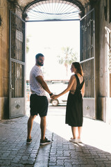 Couple of lovers is exploring new city during summer holidays. Man holds the hand of his beloved while walking down the street. A male and a female wearing casual clothes enjoy the romantic dating.