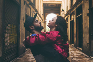 A bearded man in a cap is hugging his beloved woman while standing on a street background. Attractive woman is looking into the eyes of her husband during spending a leisure time outdoors.