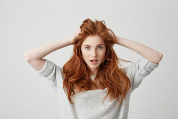 Portrait of surprised redhead beautiful girl looking at camera with opened mouth touching head over white background.