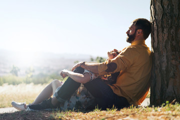 Charming couple of a man and a woman wearing casual clothes is sitting beside the tree on a natural landscape background. A man hugs a beloved woman while outdoors trip on a sunny summer day.