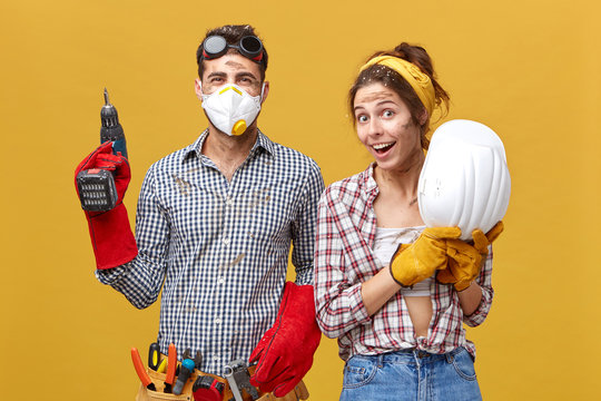 Portrait of positive male with drilling machine and tool belt and his female colleague holding hardhat having delightful expression isolated over yellow background. Manual workers and builduing