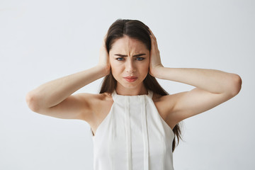 Displeased beautiful brunette girl closing ears with hands from noise over white background.