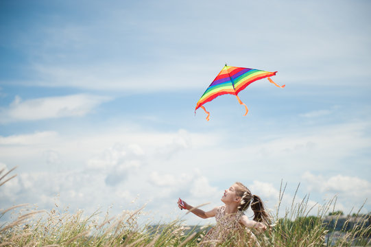 happy smiling little girl holding flying kite in field on blue cloudy sky background