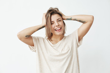 Cheerful cool tattooed girl smiling winking showing tongue looking at camera touching hair over white background.
