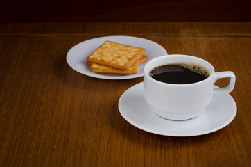 Hot black coffee in white ceramic cup on saucer over dark teak wood background served with cream crackers on white plate with copy space for text insertion