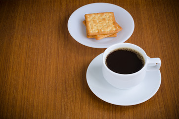 Hot black coffee in white ceramic cup on saucer over dark teak wood background served with cream crackers on white plate with copy space for text insertion